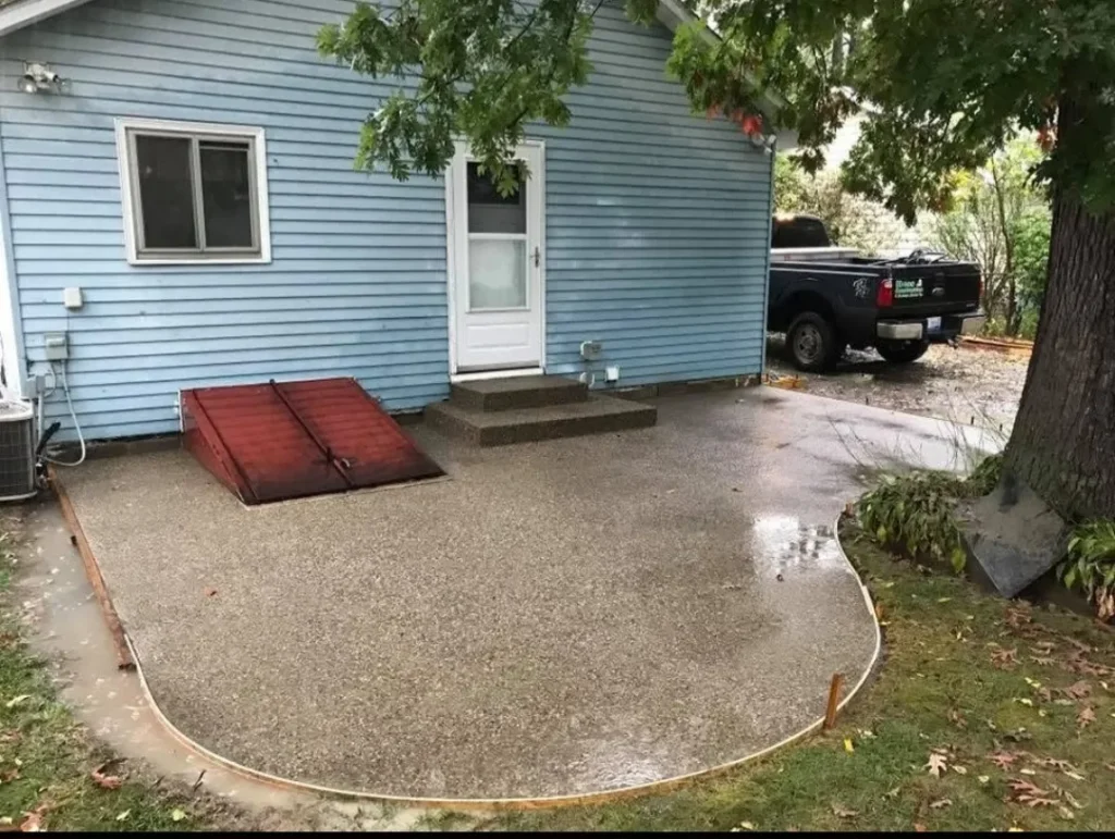 A blue house with a white door, featuring a red ramp on a rain-soaked concrete patio, surrounded by greenery and a parked black truck.