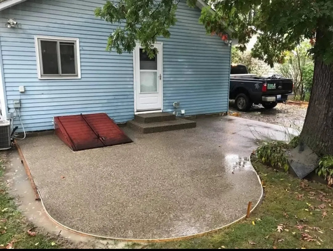 A blue house with a white door, featuring a red ramp on a rain-soaked concrete patio, surrounded by greenery and a parked black truck.