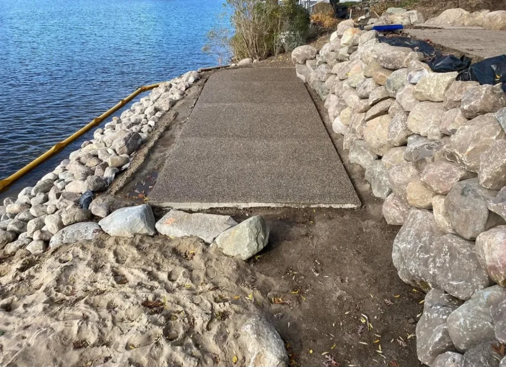 A newly constructed concrete path leads to a lake, bordered by smooth stones and sandy earth. Foliage in the background adds greenery.
