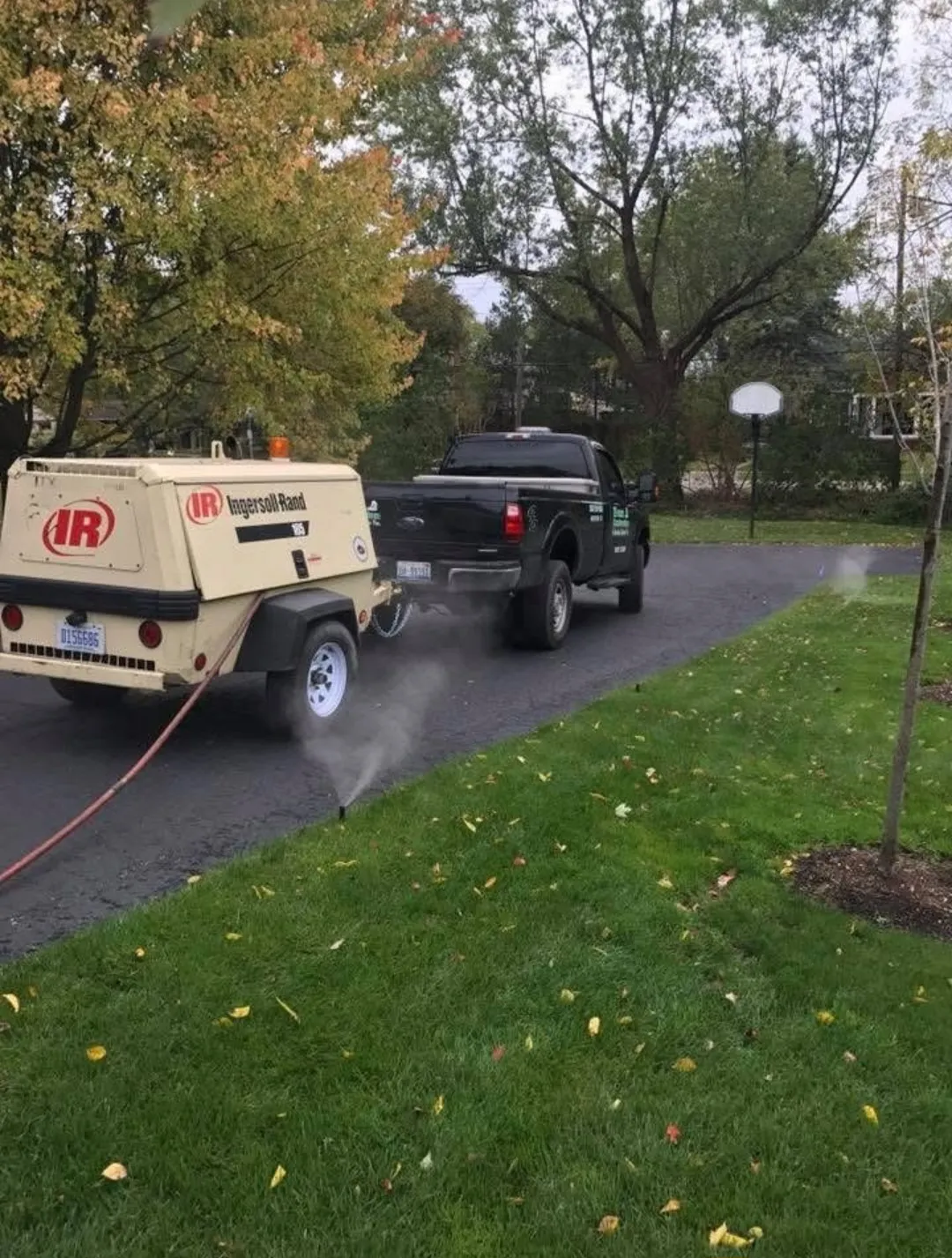 A black pickup truck towing an Ingersoll Rand air compressor along a leaf-strewn driveway, surrounded by trees and grass.