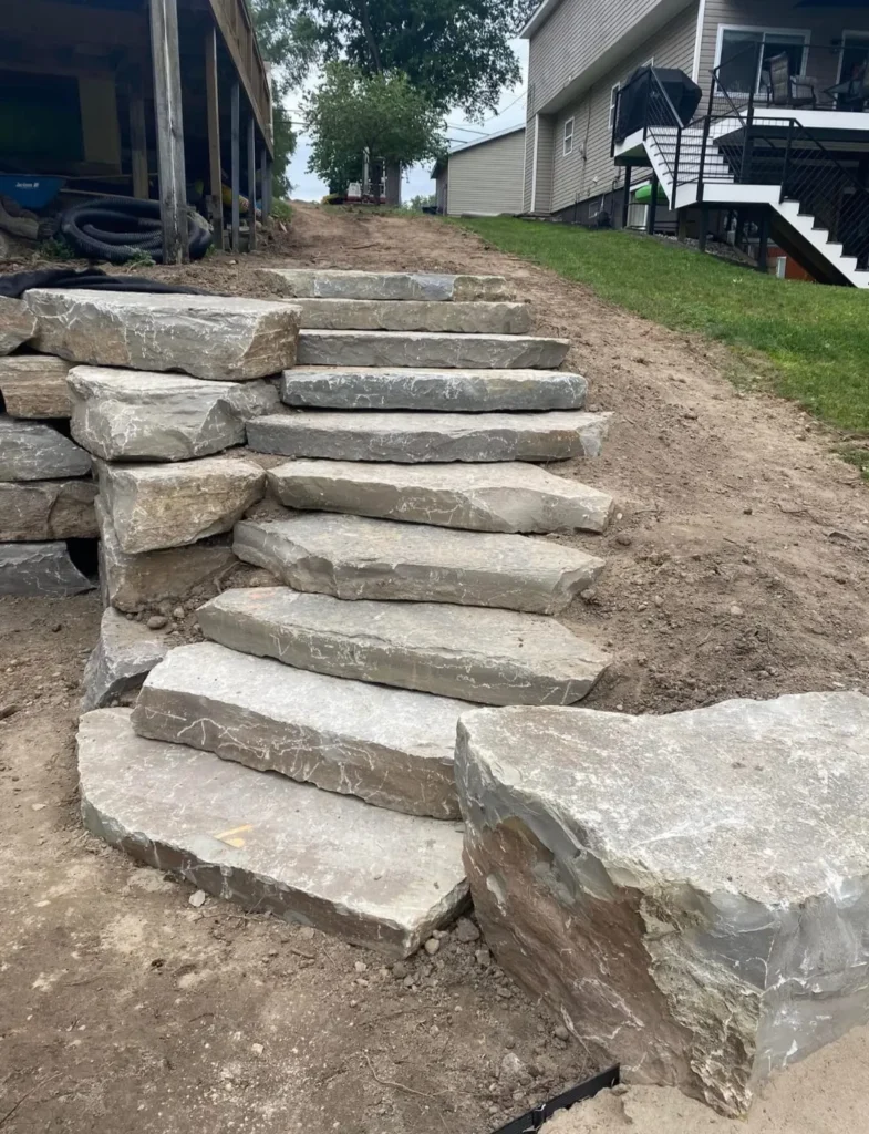 A set of natural stone steps leads up a slope in a yard, surrounded by dirt and grass, with a house visible in the background.