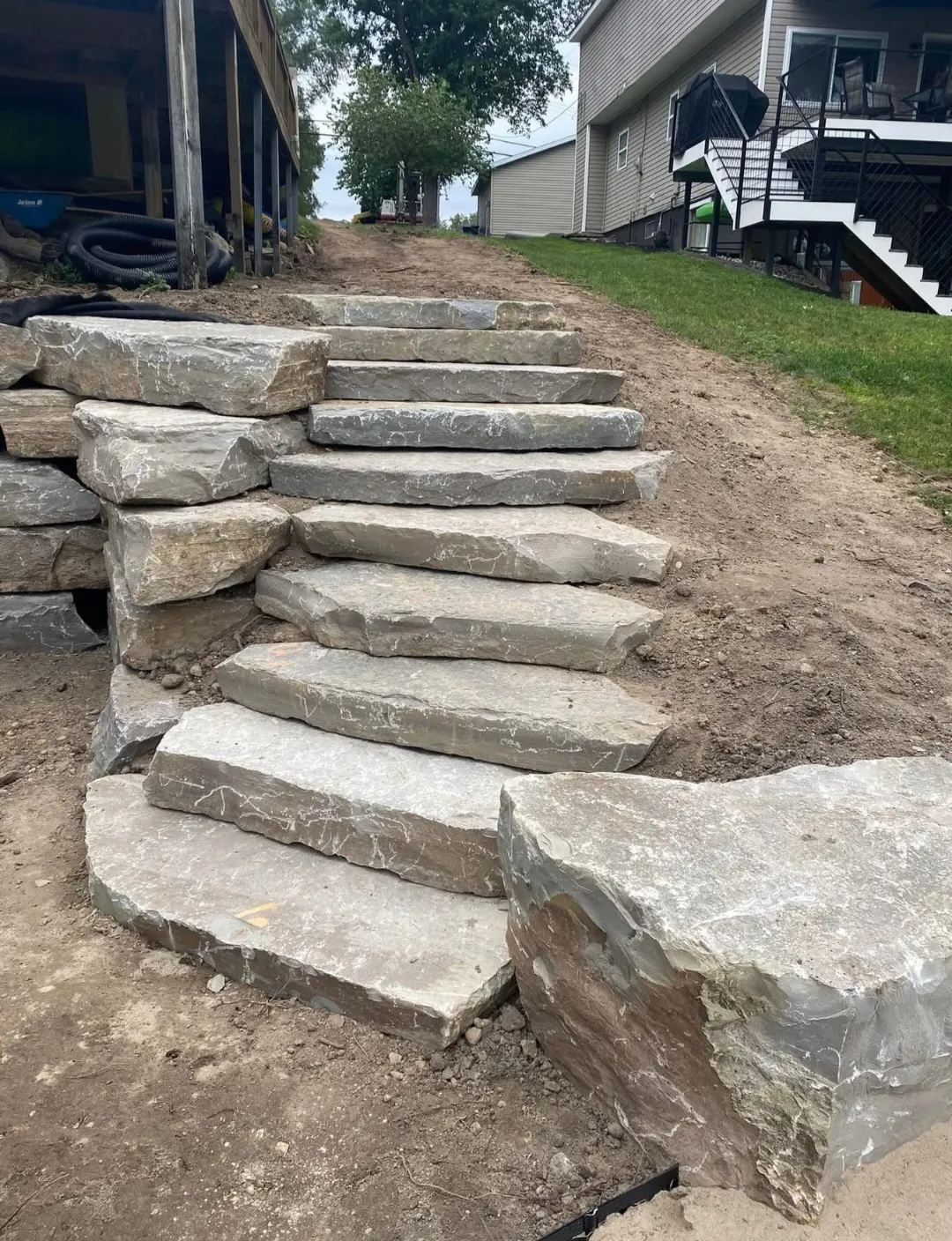 A set of natural stone steps leads up a slope in a yard, surrounded by dirt and grass, with a house visible in the background.