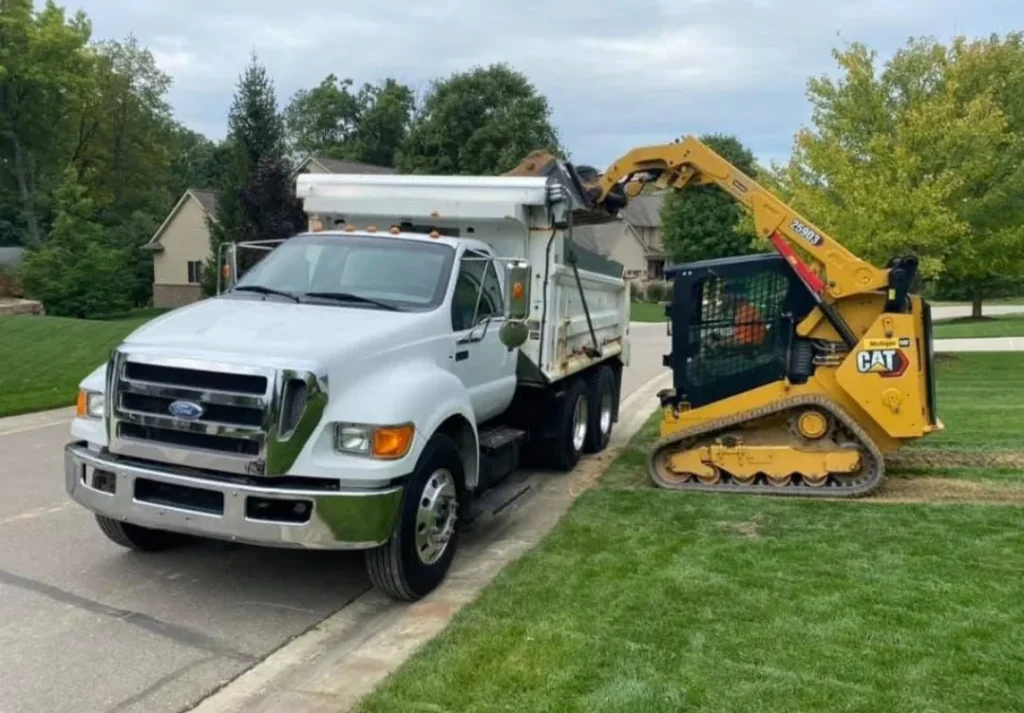 A white Ford dump truck parked on the street, with a CAT skid steer loader positioned next to it, on a grassy residential lawn.